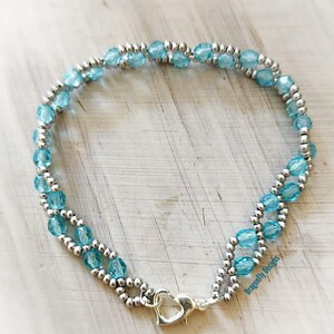 May include: A delicate bracelet with a double strand of light blue glass beads and silver beads. The bracelet has a silver heart-shaped clasp.