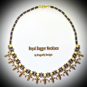 May include: A gold and purple beaded necklace with a dagger-shaped pendant. The necklace is called "Royal Dagger Necklace" by Dragonfly Designs.
