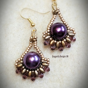 Peut inclure: Une paire de boucles d'oreilles dorées avec une perle violette et un motif de fil doré. Les boucles d'oreilles sont étiquetées "Solaris Earrings" et "Dragonfly Designs UK".