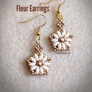May include: A pair of gold-colored Fleur earrings. Each earring features a flower design with white petals and a gold center. The earrings are suspended from gold-colored ear wires. The text "Fleur Earrings" is visible in the image.