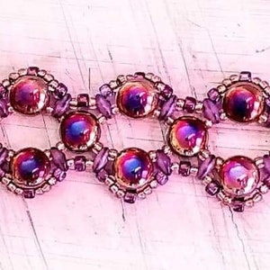 May include: A purple and copper beaded bracelet with a geometric design. The bracelet features iridescent beads and a clasp closure. "Dragonfly Designs" is printed on the bracelet.