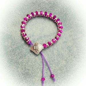 May include: A purple beaded bracelet with a silver charm and purple cord ties.