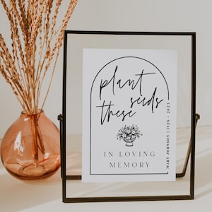 May include: A clear glass frame holding a white card with the words "plant seeds these" in black script. Below the text is a floral illustration and the words "In Loving Memory". A vase with dried flowers is to the left.