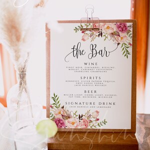 May include: A white sign with the text "the Bar" in black calligraphy. The sign is decorated with pink and green watercolour flowers and leaves. The sign lists the drinks available at the bar, including wine, spirits, beer, and a signature drink. The sign is on a wooden easel.