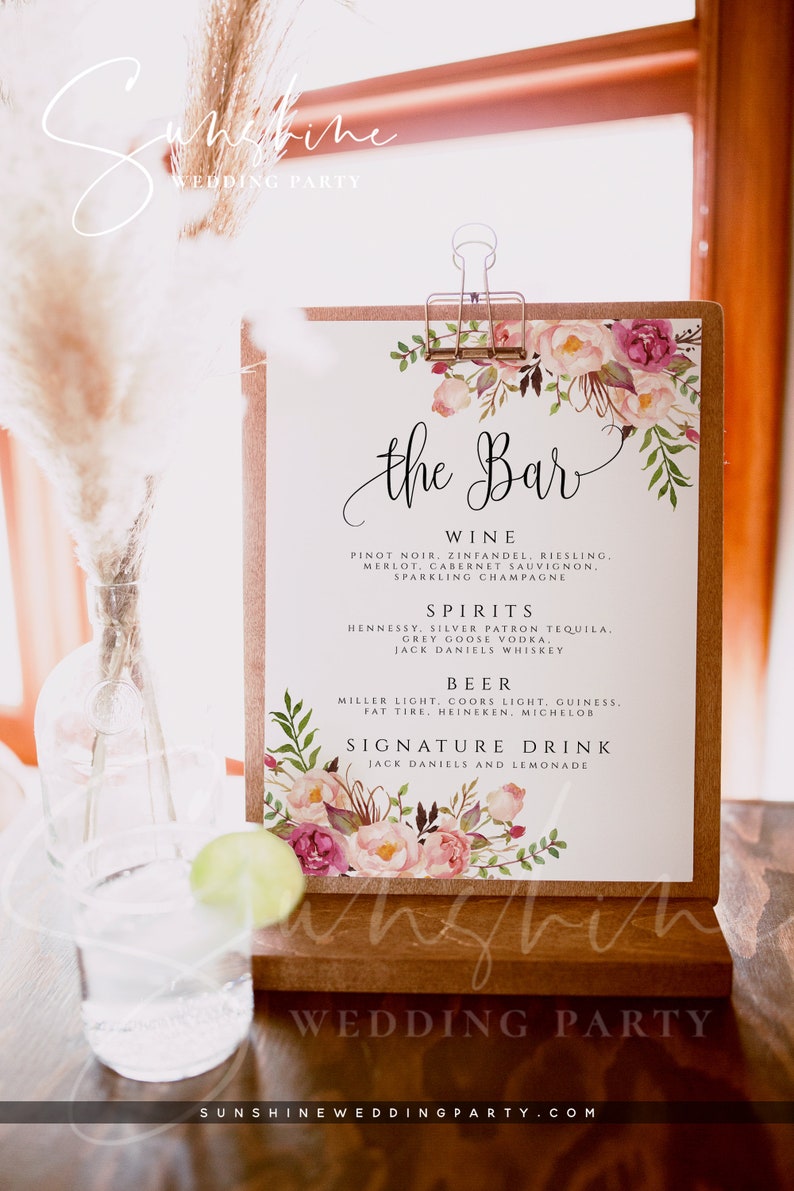 May include: A white sign with the text "the Bar" in black calligraphy. The sign is decorated with pink and green watercolour flowers and leaves. The sign lists the drinks available at the bar, including wine, spirits, beer, and a signature drink. The sign is on a wooden easel.