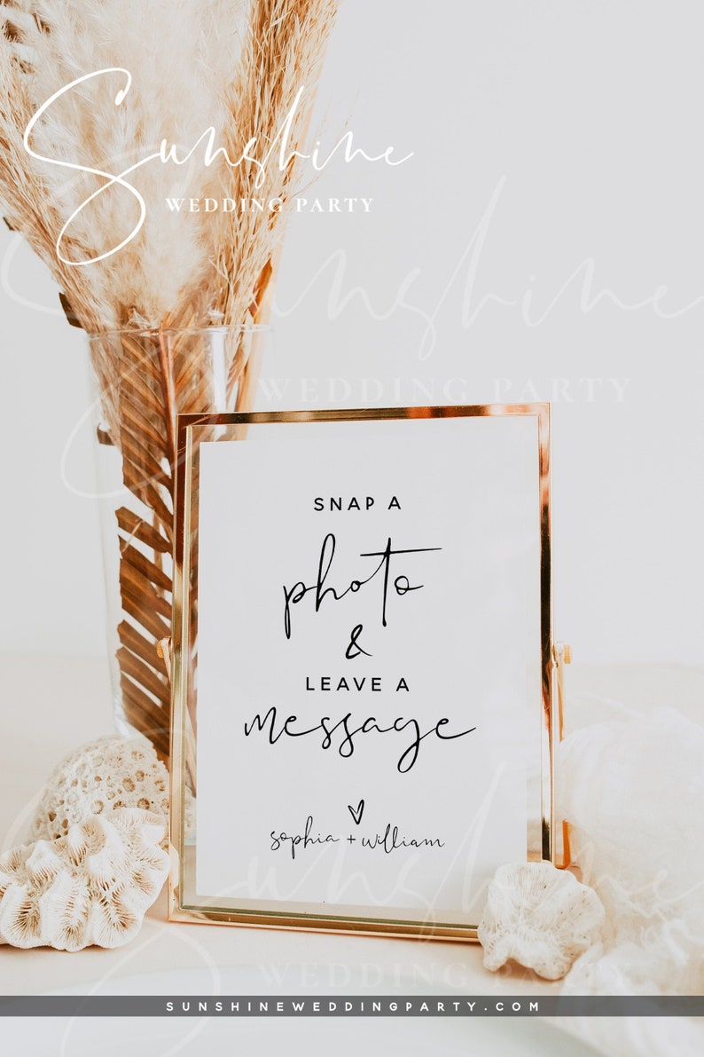 May include: A white sign with a gold frame that says "Snap a photo & leave a message" with the names "sophia + william" below. The sign is sitting on a white surface with a seashell and dried pampas grass.