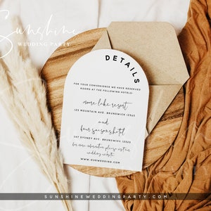 Op de afbeelding: Een witte kaart met zwarte tekst op een houten oppervlak. De kaart heeft de tekst "DETAILS" in een gebogen lettertype. De tekst op de kaart vermeldt twee hotels: "More Lake Resort" en "Four Seasons Hotel" met hun adressen. De kaart bevat ook de website "www.ourwedding.com".