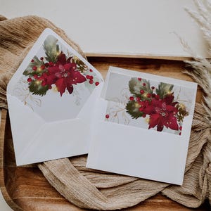 Könnte beinhalten: Zwei weiße Umschläge mit einem Weihnachtsstern-Design. Die Umschläge sind geöffnet und zeigen ein Blumenmuster mit roten Weihnachtssternen, Beeren und goldenen Akzenten. Sie liegen auf einem Holztablett mit einem braunen Tuch.