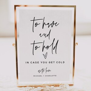 Könnte beinhalten: Ein goldfarbener Rahmen mit dem Text "to have and to hold" in schwarzer Kalligraphie. Unter dem Text befindet sich ein kleines Herz und die Worte "in case you get cold" in schwarzer Schrift. Das Schild enthält auch den Text "with love" und die Namen "Michael + Charlotte" in schwarzer Schrift.