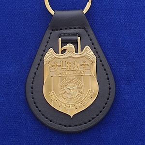 Può includere: Un portachiavi in ​​cuoio nero con un ciondolo dorato. Il portachiavi presenta una replica di un distintivo di agente speciale degli Stati Uniti con le parole "U.S. SPECIAL AGENT" e un sigillo con un'aquila.