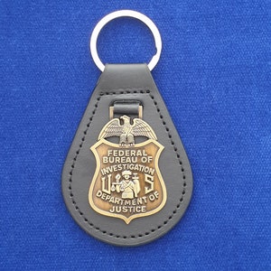 Puede incluir: Llavero de cuero negro con un emblema metálico que lleva las palabras "FEDERAL BUREAU OF INVESTIGATION" y "U.S. DEPARTMENT OF JUSTICE". El emblema tiene un acabado dorado y un diseño de águila. Un llavero plateado está adjunto.