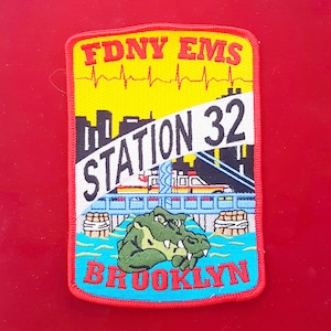 May include: A red, white and blue embroidered patch with the text "FDNY EMS Station 32 Brooklyn". The patch features a cartoon alligator and a cityscape with an ambulance driving over a bridge.