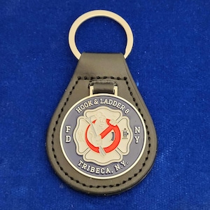 Puede incluir: Llavero de cuero negro con un dije de metal plateado que presenta un emblema de bomberos azul y rojo. El emblema dice "Hook & Ladder 8 FD NY" y "Tribeca, N.Y."