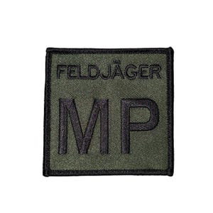 以下が含まれることがあります： 黒い刺繍文字が入った正方形のオリーブグリーンのパッチ。「FELDJÄGER」が上段に、「MP」が下段に書かれています。パッチには黒い縁取りがあります。