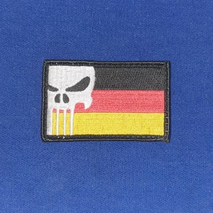 Puede incluir: Un parche bordado en blanco y negro con un diseño de calavera y huesos cruzados sobre una bandera alemana. La bandera es negra, roja y amarilla.