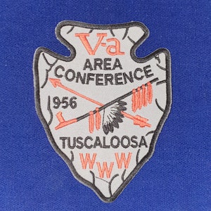 Op de afbeelding: Een witte patch in de vorm van een pijlpunt met de tekst "Va Area Conference 1956 Tuscaloosa WWW" in zwarte en roze letters.