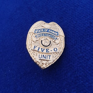 Può includere: Un distintivo color oro con la scritta "STATE OF HAWAII INVESTIGATOR" e "FIVE-O UNIT" in lettere blu e nere. Il distintivo ha una forma a scudo con un design dettagliato su sfondo blu.