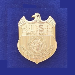 May include: A gold-colored badge with the text "U.S. SPECIAL AGENT" and the seal of the U.S. Department of Homeland Security, Naval Criminal Investigative Service.