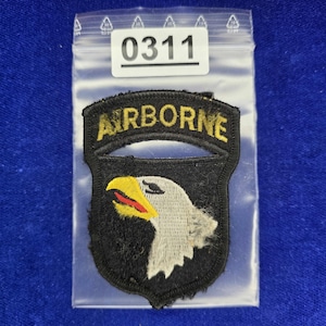 Puede incluir: Un parche bordado negro con la palabra AIRBORNE en letras amarillas sobre una cabeza de águila blanca con pico amarillo y lengua roja. El parche está dentro de una bolsa de plástico transparente con una etiqueta blanca con el número 0311.