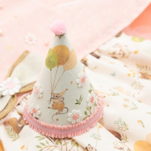May include: A white and pink party hat with a pink pom-pom on top. The hat has a mouse with balloons and flowers printed on it.