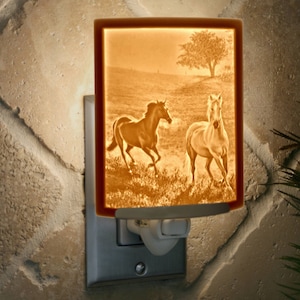 May include: A ceramic night light with a picture of two horses running in a field. The night light is plugged into an outlet and is lit up.