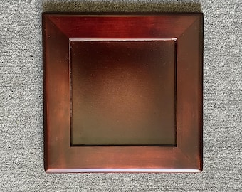 May include: A square, wooden display stand with a rich, dark cherry finish. The stand features a recessed center panel and a beveled edge, creating a framed appearance. The stand is designed to showcase decorative items.