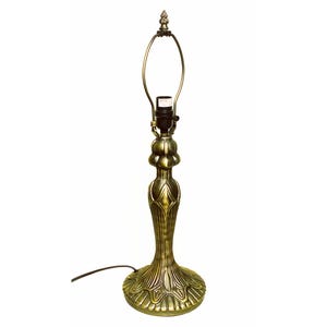 May include: A brass-toned metal table lamp base with a flared, ribbed circular pedestal and a central column featuring organic, petal-like detailing. The lamp includes a black socket and a tall, oval-shaped metal harp with a decorative finial.