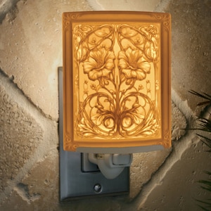 Puede incluir: Una luz de noche decorativa con un diseño floral. La luz rectangular presenta un relieve detallado de dos flores y follaje dentro de un marco dorado. La luz de noche está enchufada a una toma de corriente y emite un brillo cálido y suave.