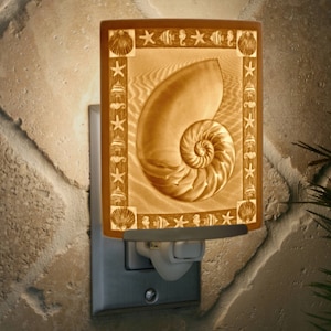 May include: A ceramic night light featuring a seashell design. The seashell is a nautilus shell with a spiral pattern. The night light is illuminated and has a white cord.