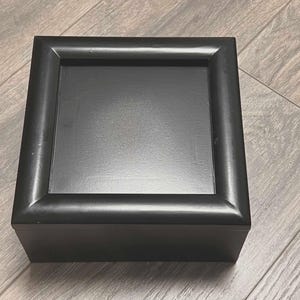 May include: A square, black wooden box with a recessed top. The box has a simple, elegant design with clean lines and a matte finish. The box is sitting on a wooden surface.