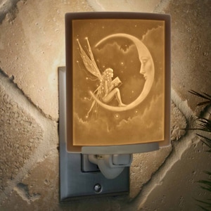 May include: A ceramic night light with a fairy sitting on a crescent moon reading a book. The night light is illuminated and has a white background with a beige crescent moon and fairy.