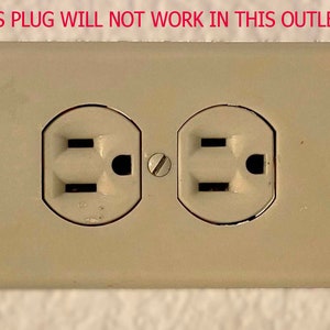 Op de afbeelding: Een close-up van een beige stopcontact met twee standaard stopcontacten met drie pinnen. De tekst "THIS PLUG WILL NOT WORK IN THIS OUTLET" staat boven het stopcontact.