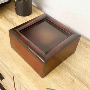May include: A rectangular wooden box with a dark brown finish. The box has a raised, framed lid with a recessed center. The wood grain is visible, and the box appears to be well-crafted. The box is sitting on a wooden surface.