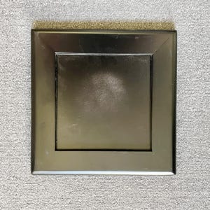 May include: A square, dark bronze-colored metal vent cover. The cover has a recessed center and a raised, beveled frame. The surface reflects light, creating highlights and shadows. The vent cover is likely for a floor or wall.