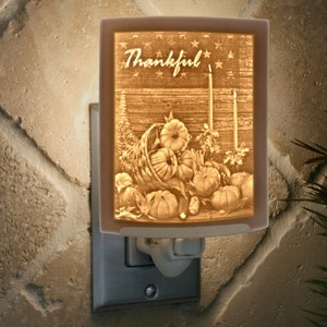 May include: A ceramic night light with a Thanksgiving theme. The image features a cornucopia overflowing with pumpkins and the word "Thankful" on a rustic wooden background.