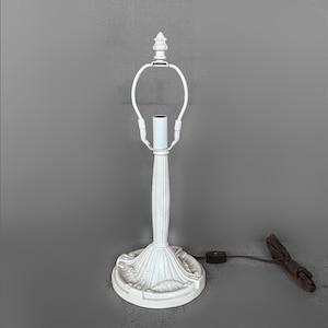 May include: A white table lamp base featuring a fluted central column and a decorative, textured circular pedestal. The lamp includes a metal harp assembly for shade attachment, a central light socket, and a brown electrical cord with a toggle switch.
