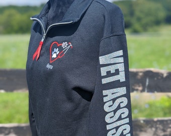 May include: Black fleece pullover with a zipper and a red heart with a paw print and stethoscope design on the front. The text "Vet Assistant" is printed in silver glitter on the left sleeve.