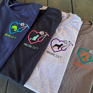 May include: Four t-shirts with different colored embroidered hearts featuring a stethoscope, paw print, and animal silhouette. The text "Nicole, LVT" is embroidered on each shirt.