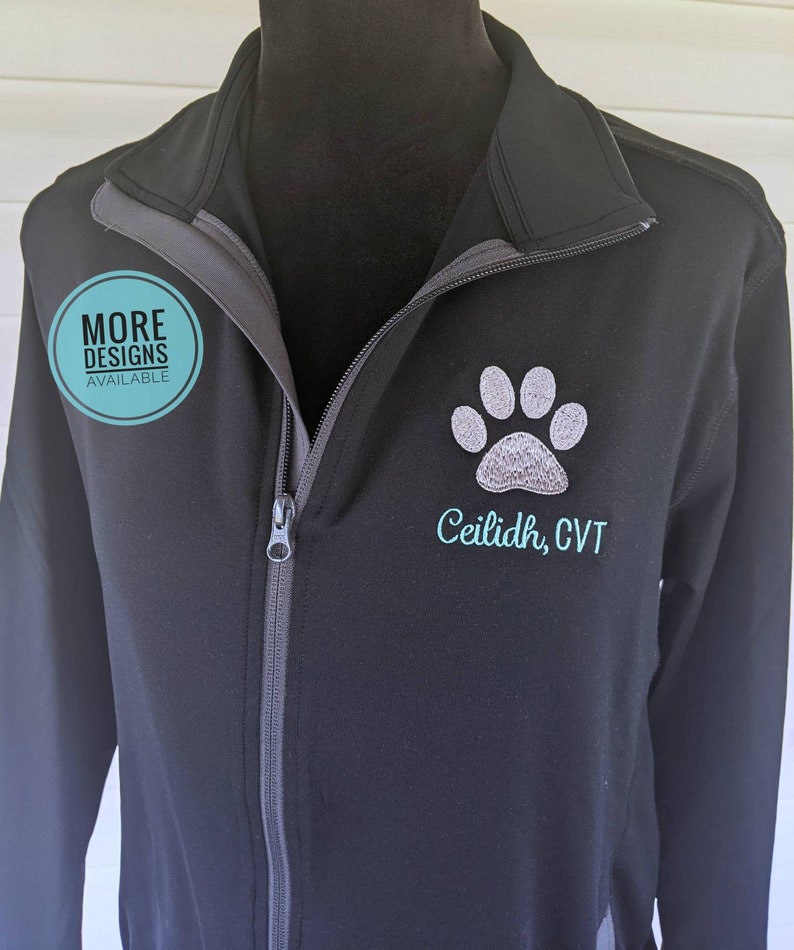 May include: Black zip-up jacket with a gray paw print design and the name "Ceilidh, CVT" embroidered in teal. The jacket has a high collar and a full-length zipper. A round teal sticker says "MORE DESIGNS AVAILABLE".