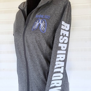 May include: A gray zippered jacket with a white embroidered lung design and the text "Amber RRT" on the left chest. The right sleeve has the word "RESPIRATORY" printed vertically in white.