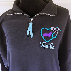 May include: Black sweatshirt with a quarter-zip collar and a light blue ribbon on the zipper. It has an embroidered design of a purple corgi within a blue heart, a paw print, a stethoscope, and the name "Kaitlin" in script.