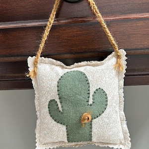 Puede incluir: Un cojín cuadrado decorativo con un estampado floral beige claro y una aplicación de cactus verde. El cojín tiene un borde festoneado y cuelga de una cuerda marrón. Un botón de madera está cosido en el cactus.