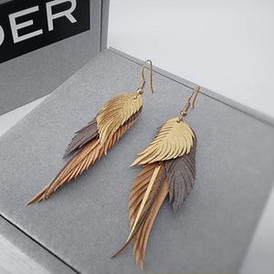 Könnte beinhalten: Ein Paar goldene und braune Lederfederohrringe. Die Ohrringe sind lang und hängen herunter, wobei die Federn übereinander gelegt sind.