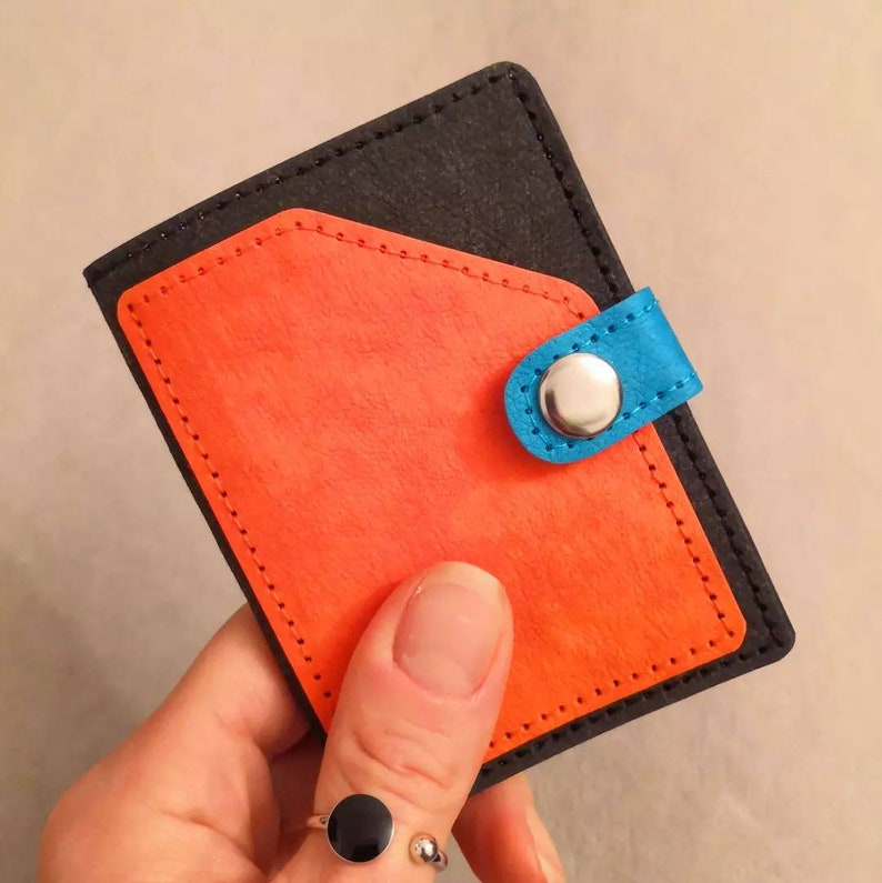 May include: A small, black leather wallet with a bright orange flap and a blue snap closure. The wallet has a stitched edge and a silver button.