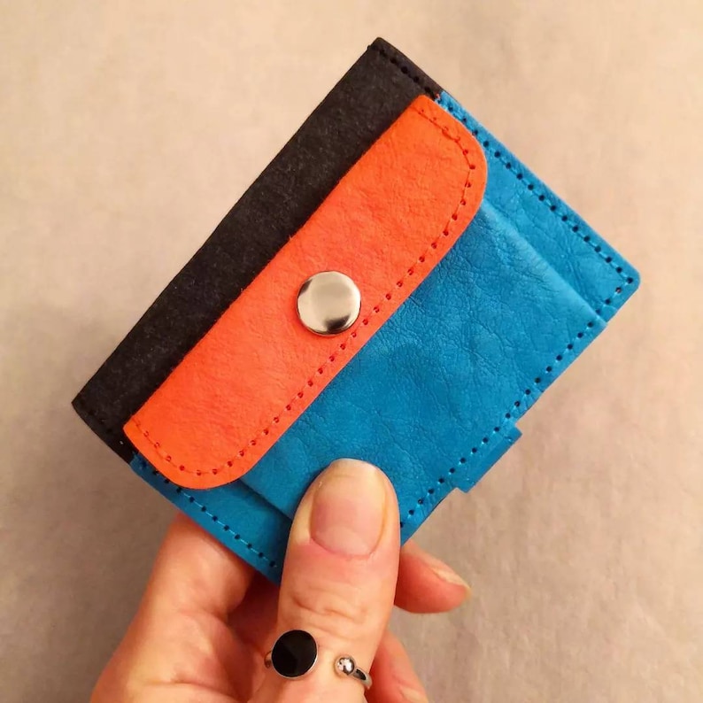 May include: A small, blue leather wallet with a flap that has a bright orange lining and a black felt backing. The wallet has a silver snap closure.