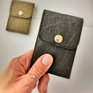 May include: Two small, rectangular cases with a flap and snap closure. One case is olive green, the other is dark gray. The cases have a textured appearance and a silver-colored snap. The cases are likely designed to hold small items.