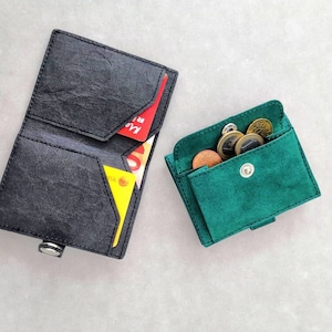 May include: Two leather wallets, one black and one green. The black wallet has a fold-over design and is open to reveal three cards. The green wallet is closed and has a flap with a snap closure. Both wallets have a textured surface.