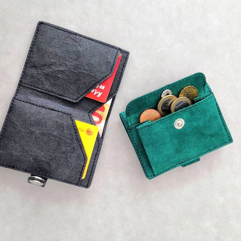May include: Two leather wallets, one black and one green. The black wallet has a fold-over design and is open to reveal three cards. The green wallet is closed and has a flap with a snap closure. Both wallets have a textured surface.
