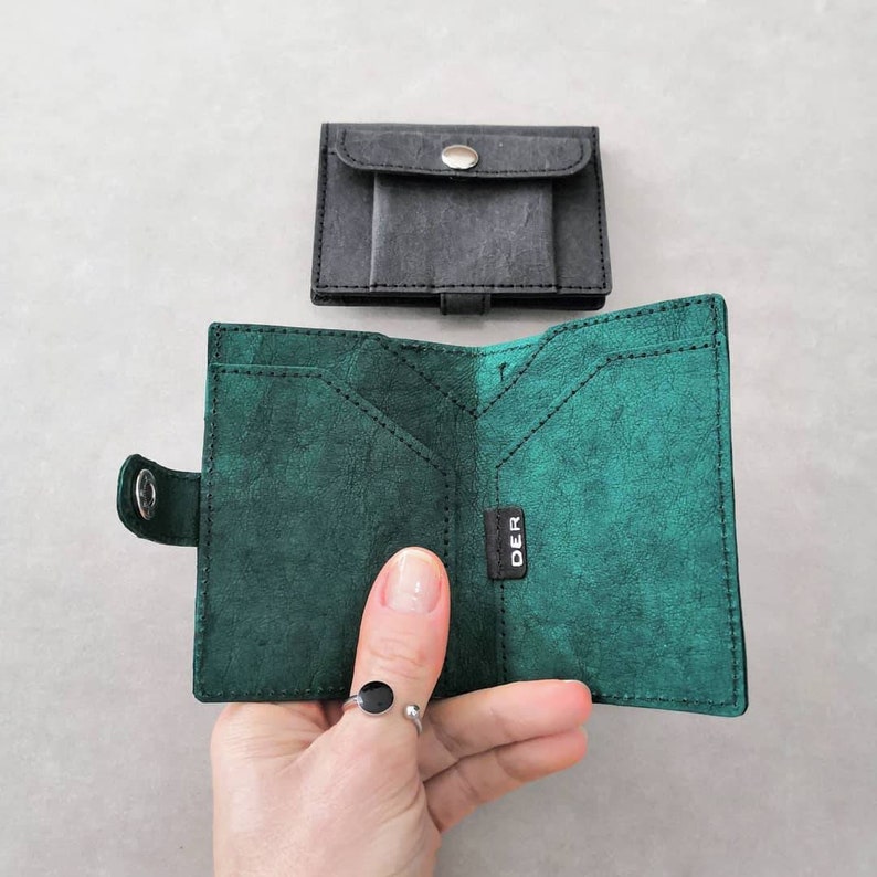 May include: A green leather wallet with a black leather flap and a silver button. The wallet is open and has multiple card slots and a money pocket. The brand name "DER" is visible on the inside of the wallet.