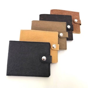May include: A collection of five rectangular wallets arranged in a staggered row. Each wallet features a textured surface, a single snap button closure, and visible stitching along the edges. The colors include black, tan, olive, dark brown, and light brown.